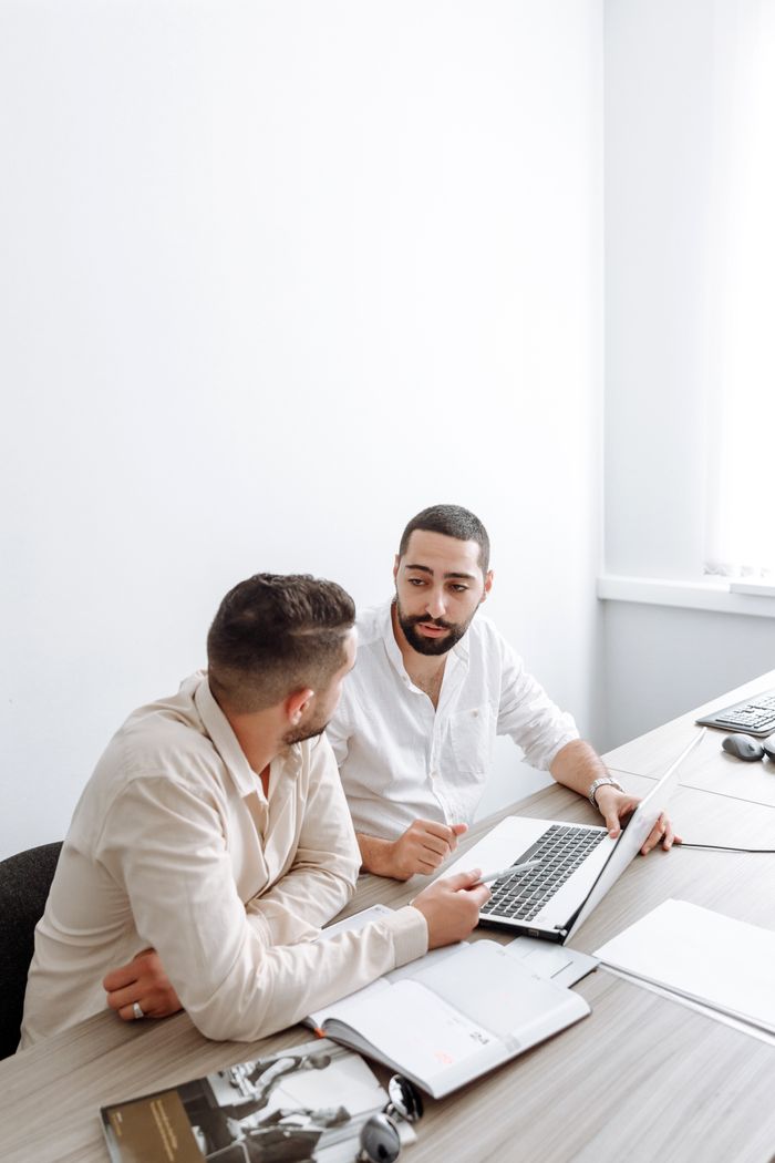 about-us Two businessmen in white shirts engaged in a discussion over a laptop in a bright office setting.