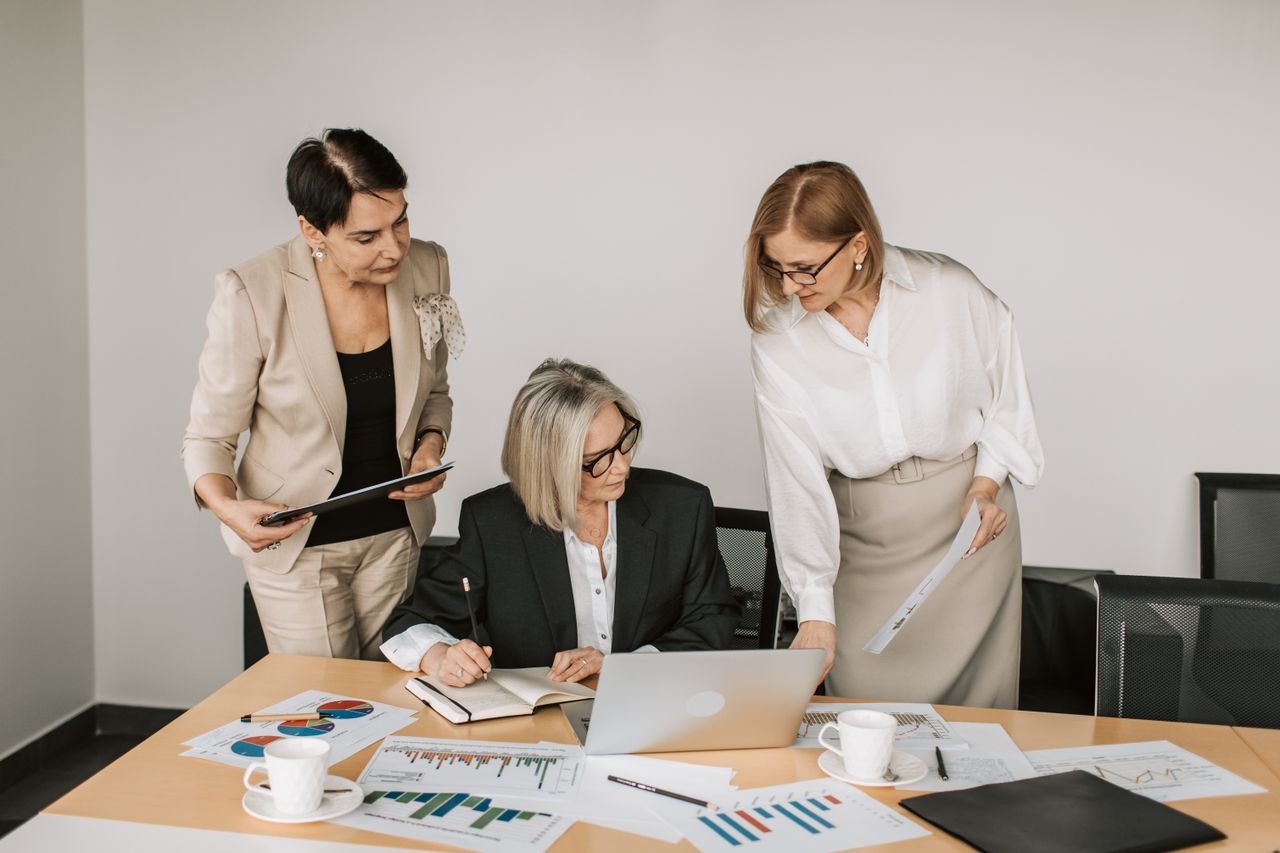 who-we-are Three businesswomen discussing documents during a meeting in a modern office setting.